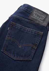 Tmavě modré džínové kalhoty složené tak, aby byla vidět zadní kapsa s oranžovým prošíváním a koženým štítkem značky Levi Strauss & Co. nad kapsou.
