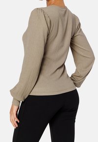 Donna che indossa una blusa lunga beige a maniche lunghe con spalle a palloncino e pantaloni neri aderenti, in posa con una mano sui fianchi.