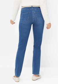 Jean en denim bleu avec une coupe droite, taille mi-haute, deux poches arrière et une texture lisse. Porté avec un pull crème et des chaussures claires.