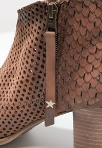 Botín de tobillo de cuero perforado marrón con un diseño ondulado, una cremallera lateral y un tirador de cuero con un acento de estrella plateada.