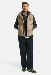 Beige gesteppte Weste mit Seitentaschen und einem braunen Logo-Patch, getragen über einem dunkelgrünen Pullover, kombiniert mit navyblauen Hosen und grünen Sneakers.