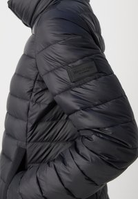 Veste rembourrée noire avec un design matelassé, dotée d'un col montant et d'un panneau latéral. Inclut un écusson en relief sur la manche.