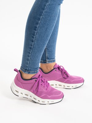Person trägt blaue Jeans und leuchtend pinke Sportschuhe mit dicken weißen Sohlen, steht auf einer weißen Oberfläche.