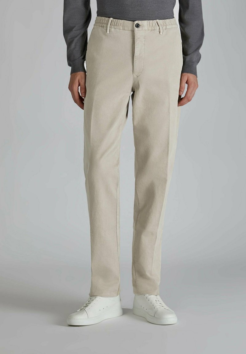 Homme portant un pantalon beige à jambes droites, des baskets blanches et une chemise grise à manches longues, debout devant un fond gris uni.