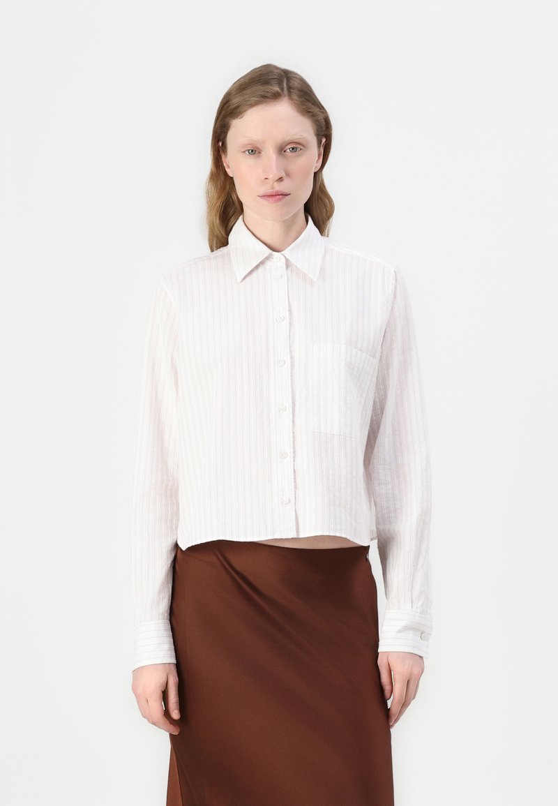Camicia bianca a maniche lunghe con bottoni, caratterizzata da righe verticali, una tasca sul petto e un orlo accorciato. Indossata con una gonna in satin marrone.