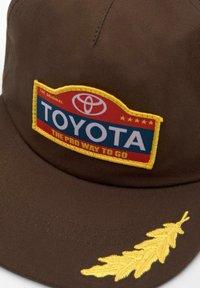 Brązowa czapka z naszywką w niebiesko-czerwonym logo Toyoty z żółtymi akcentami i tekstem, a także wyszywaną żółtą haftką liścia z boku.