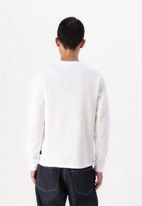 Langärmliges weißes Hemd aus strukturiertem Stoff, mit Rundhalsausschnitt und gerippten Bündchen, kombiniert mit dunkelblauen Jeans.