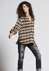 Langärmlige Bluse aus gemustertem Stoff mit schwarzen und goldenen ovalen Formen, kombiniert mit zerrissenen grauen Jeans, die Risse und Patches aufweisen.