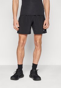 Schwarze Sportshorts mit glatter Textur und elastischem Bund. Getragen mit schwarzen Laufschuhen und Socken in Knöchellänge.