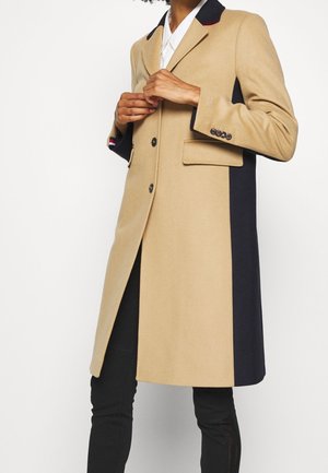 Osoba nosząca długi płaszcz w beżowo-czarne bloki kolorów, zapinający go z przodu, w połączeniu z czarnymi spodniami i białą koszulą pod spodem.