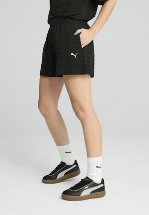 Czarne krótkie spodenki w paski z elastycznym pasem, bocznymi kieszeniami i białym logo; zestawione z czarnymi sneakersami i białymi skarpetkami z prążkami.
