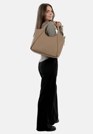 Borsa a mano in finta pelle beige con un design angolare, due manici a spalla e una tasca con zip. Il modello indossa una maglietta grigia e pantaloni neri.