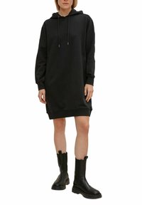 Personne portant une robe sweat à capuche noire longue associée à des bottes épaisses noires mi-mollet, debout devant un fond uni.
