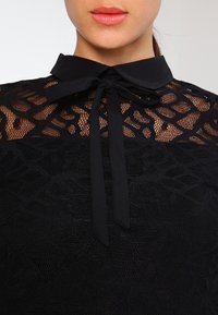 Sort bluse med høj krave og bundet sløjfe. Teksturdesign af blonde præsenterer indviklede mønstre. Blødt stof med gennemsigtige sektioner er synlige.