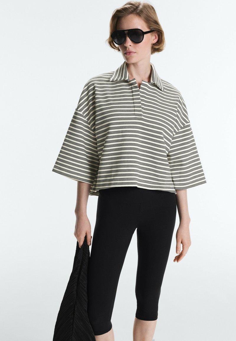 Femme portant de grandes lunettes de soleil noires, chemise ample rayée à manches larges, leggings noirs courts, tenant un tissu noir texturé.