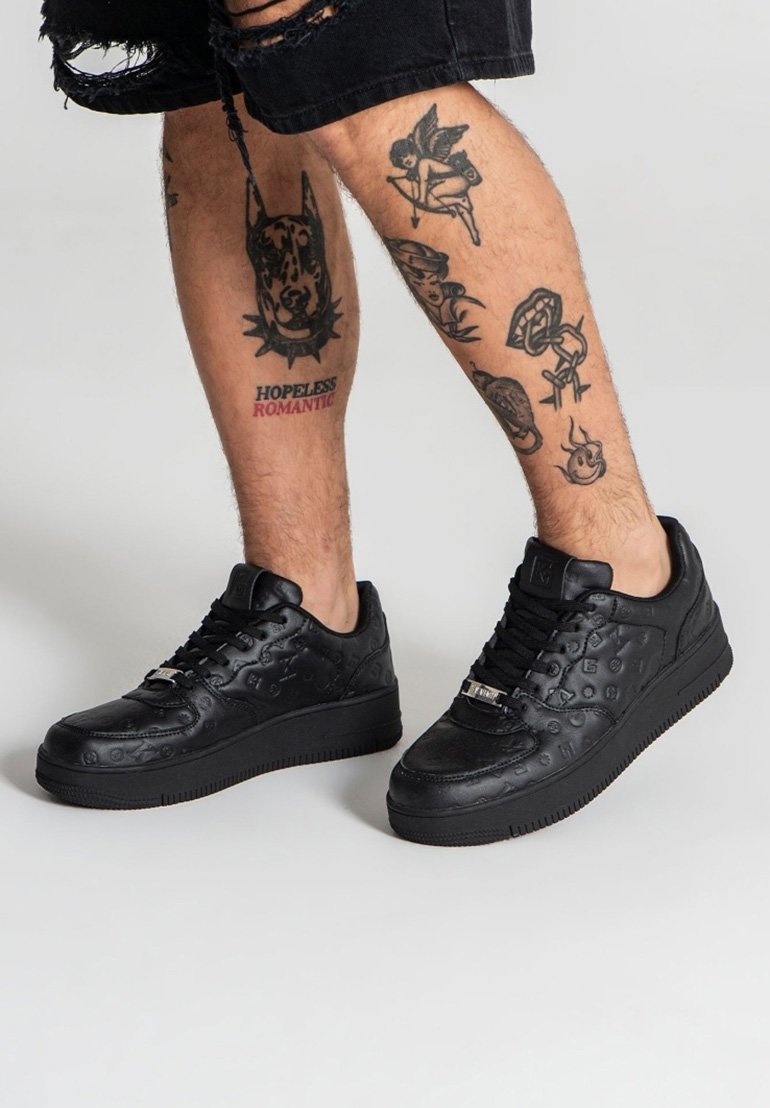 Sneaker nere in pelle texturizzata con loghi in rilievo, dotate di lacci piatti e suola in gomma spessa. Tatuaggi visibili sulle gambe.