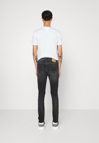 Jeans de mezclilla negra con corte ajustado, que presentan un parche de cuero marrón en la cintura trasera y un sutil desgaste para añadir textura. Combinados con una camiseta blanca.