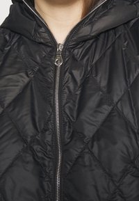 Černá prošívaná vesta s kapucí, která má přední zip, diamantový vzor z látky a hladký povrch.