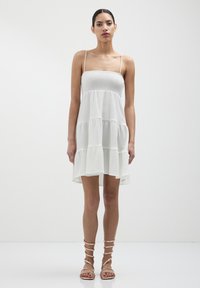 Abito estivo bianco a più strati con spalline sottili, tessuto testurizzato e una vestibilità rilassata, abbinato a sandali con cinturini. Design minimale.