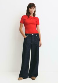 Rood aansluitend T-shirt met een geplooide halslijn, gecombineerd met donkerblauwe denim jeans met brede pijpen, voorzakken en contrasterende stiksel details.