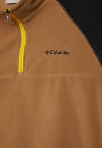 Ljust brunt fleece-pullov med en gul halvdragkedja vid kragen. Svart axeldetaljering och en svart Columbia-logotyp på bröstet. Mjukt material.