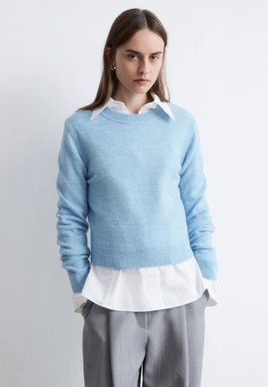 Hellblauer Strickpullover mit rundem Ausschnitt, gerippten Bündchen und Saum, darüber getragen ein weißes Hemd mit sichtbarem Kragen und Manschetten. Graue Hose.