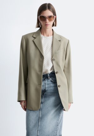 Kvinde iført oversized khaki blazer, hvidt stribet top, denimnederdel, store hoop-øreringe og oversized brune solbriller mod ensfarvet baggrund.