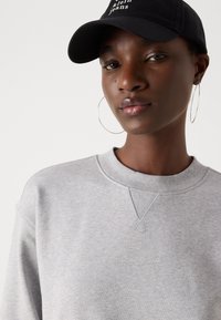 Felpa grigia con scollatura tonda e cuciture strutturate, abbinata a un cappellino nero con un marchio bianco ben visibile e grandi orecchini a cerchio.