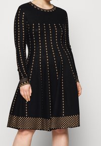 Robe noire avec motifs géométriques dorés, manches longues, encolure ronde et jupe évasée. Tissu texturé avec une finition lisse.