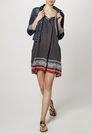 Black patterned dress with a flared shape, layered with a denim jacket. Features a red and blue hem and gold sandals.