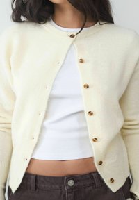 Cremfarbener Cardigan aus weichem Material, mit rundem Hals, sieben Holzknöpfen und gerippten Bündchen. Getragen über einem weißen Oberteil.