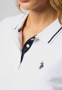 Záber zblízka osoby nosiacej biele polo tričko s tmavomodrým lemom na golieri, gombíkmi a malým vyšívaným logom hráča polo na hrudi.