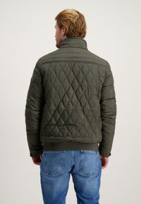 Olijfgroene gewatteerde pufferjack met een hoge kraag, ribbelboord en gestructureerde diamantpatronen op de rug. Gedragen over een blauwe spijkerbroek.