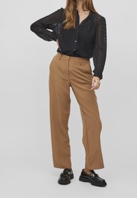 Chemisier en dentelle noire à boutons avec des manches longues, associé à un pantalon marron taille haute à jambes larges et des mocassins noirs épais.