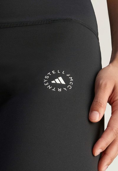Crne atletske tajice napravljene od glatkog materijala, s bijelim kružnim logotipom s tri pruge i natpisom "Stella McCartney" na butini.