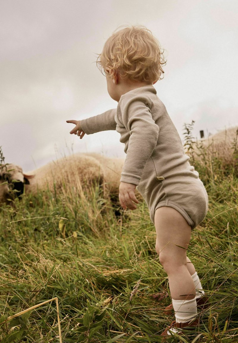 Jeune enfant aux cheveux blonds bouclés, vêtu d'une tenue beige, debout dans l'herbe haute, pointant des moutons qui broutent à proximité.