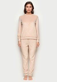 Beżowy zestaw odzieży domowej z teksturowanej tkaniny w falisty wzór. Bluzka z długim rękawem i elastyczne spodnie z mankietami, luźny krój, bez widocznych elementów metalowych.