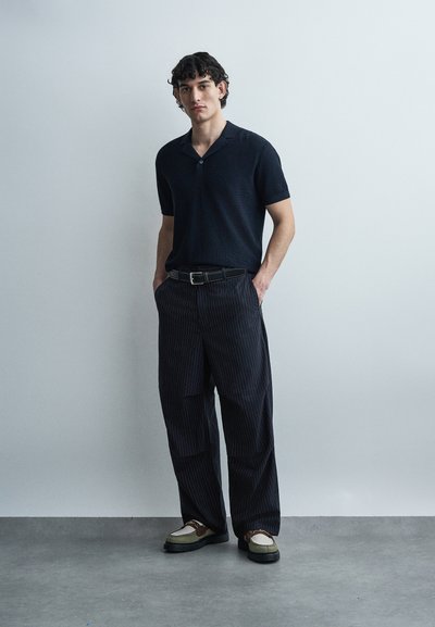 Νεαρός άνδρας στέκεται μπροστά σε έναν απλό τοίχο, φορώντας μαύρο κοντομάνικο πουκάμισο, παντελόνια με ρίγες και φαρδιά μπατζάκια, καθώς και δίχρωμα μοκασίνια.