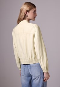 Ljusgul bomberjacka med elastisk midja och ärmslut, mjuk textur, med en enkel design och ett bakstycke med söm, som bärs över blå jeans.