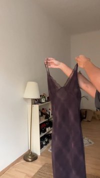 Fialové hedvábné šaty držené v rukou; mají tenká ramínka a krajkový lem. Poloprůhledná textura s hladkým, plynulým tvarem.