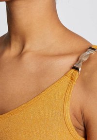 Närbild av ett gult, skimrande tyg med en texturerad yta. Remmen har en metallisk detalj med en intrikat vävd design.