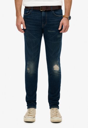Mand iført mørkeblå slidte jeans med falmede pletter på knæene, beige sneakers, brun bælte og armbåndsur, stående med afslappede hænder.