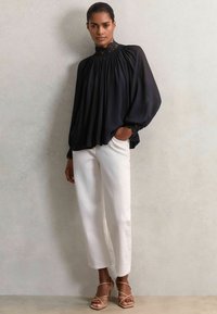 Schwarze Bluse mit hochgeschlossenem Kragen, gerafftem Stoff und Spitze, kombiniert mit weißen geraden Hosen und beigen Riemchenheels.