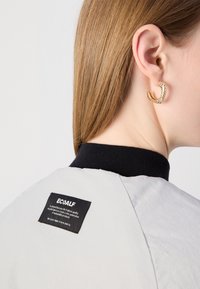 Grå jakke med svart ribbet krage. Lappen viser "ECOALF." Gull hoopsørering med teksturert overflate brukt på øret.