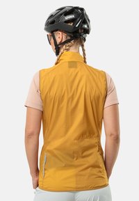 Geel lichtgewicht vest met hoge kraag en ritszakken, gecombineerd met een lichtroze shirt met korte mouwen. Zwart helm en zonnebril gedragen.