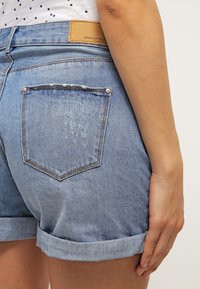 Shorts de denim en azul claro con dobladillo enrollado, que cuentan con un bolsillo trasero con ligero desgaste y una etiqueta de marca de cuero en la cinturilla.