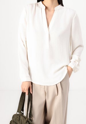 Personne portant un chemisier blanc à manches longues et un pantalon beige, tenant un sac à main texturé vert foncé dans une main, l'autre main dans la poche.