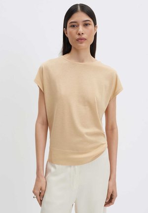 Femme aux longs cheveux foncés portant un haut beige sans manches et un pantalon clair, debout devant un fond clair uni.