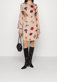 Růžové saténové šaty s barevnými potisky hub, ovoce a abstraktních tvarů. Doplněné o vysoké černé boty s špičatou špičkou a malou kabelku.