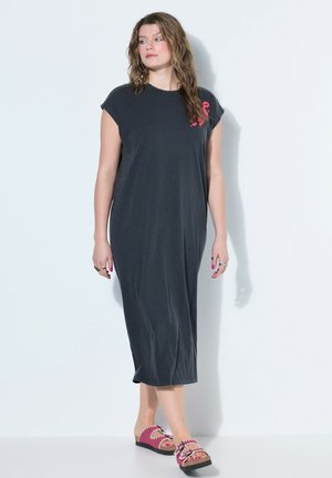 Kvinde med bølget hår iført en sort ærmeløs midikjole og pink platformsandaler står op ad en hvid væg.